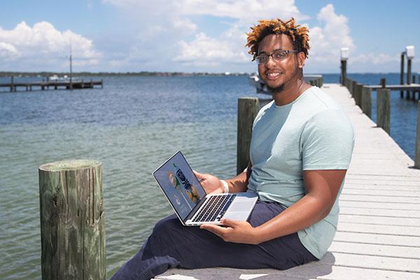 A UWF student smiles while sitting on a dock and using a laptop.