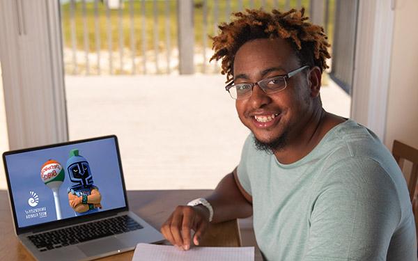 A UWF student smiles and writes with pen and paper by a laptop at home.