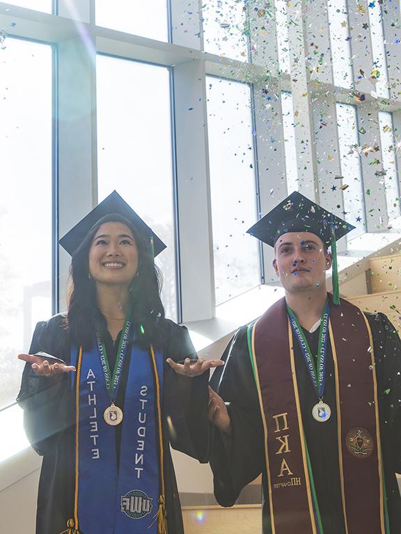 Two UWF graduates in cap and gown attire throwing confetti