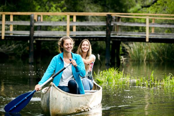 Two UWF students paddling down the river in a canoe with a walkway behind them.