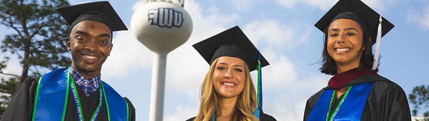 Three graduates smile while wearing cap and gown in front of the UWF water tower.